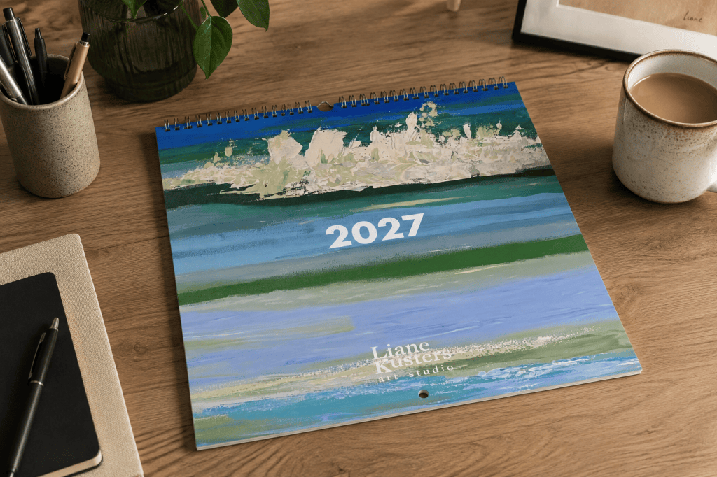 Calendar 2027 by Liane Kusters displayed closed on a wooden desk, featuring an abstract seascape painting on the cover with a metal wire binding and hanging hole.