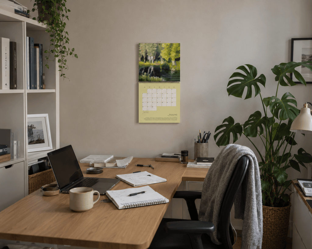 Calendar 2027 by Liane Kusters hanging open in a home office, displaying the June artwork and calendar page above a working desk.